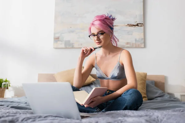 Mujer joven con el pelo teñido sosteniendo pluma y cuaderno mientras estudia en línea en casa - foto de stock