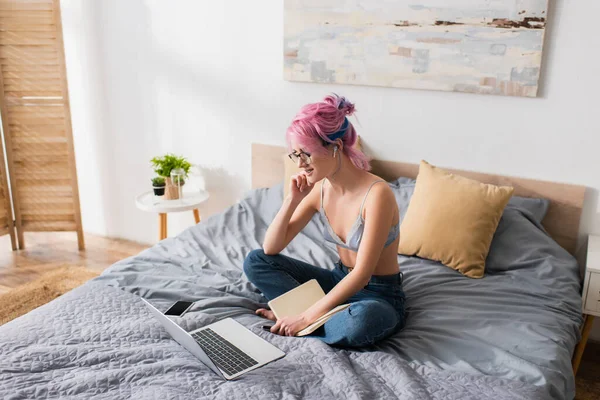 Allegra giovane donna con i capelli tinti ascolto lezione online in auricolare vicino gadget sul letto — Foto stock