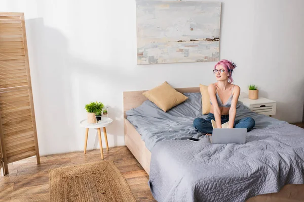 Heureuse jeune femme avec les cheveux teints écoute leçon en ligne dans écouteur près de gadgets sur le lit — Photo de stock