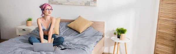 Mujer joven feliz con el pelo teñido escuchar lección en línea en auriculares cerca de gadgets en la cama, pancarta - foto de stock