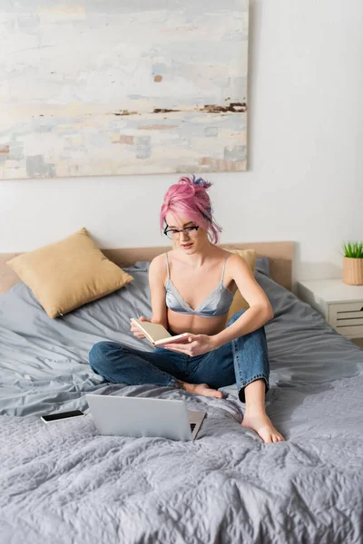 Junge Frau mit rosa Haaren hält Notizbuch während des Online-Studiums in der Nähe von Gadgets auf dem Bett — Stockfoto