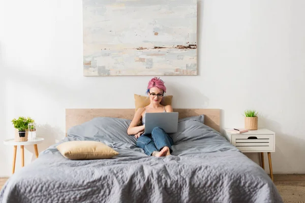 Lächelnde Frau mit gefärbten Haaren, die in BH und Jeans liegt, während sie sich den Film auf dem Laptop ansieht — Stockfoto