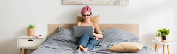 Mujer sonriente con el pelo teñido acostado en el sujetador y los pantalones vaqueros mientras mira la película en el ordenador portátil - foto de stock
