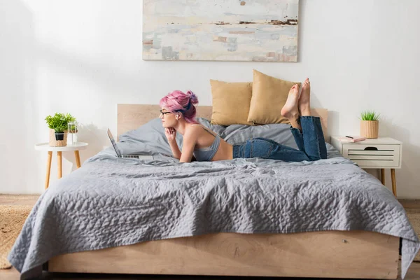 Vue latérale de femme pieds nus avec cheveux teints et écouteur couché sur le lit et regarder le film sur ordinateur portable — Photo de stock