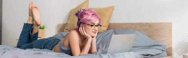 Босоногая женщина с окрашенными волосами, лежащая на кровати и смотрящая фильм на ноутбуке, баннер — стоковое фото