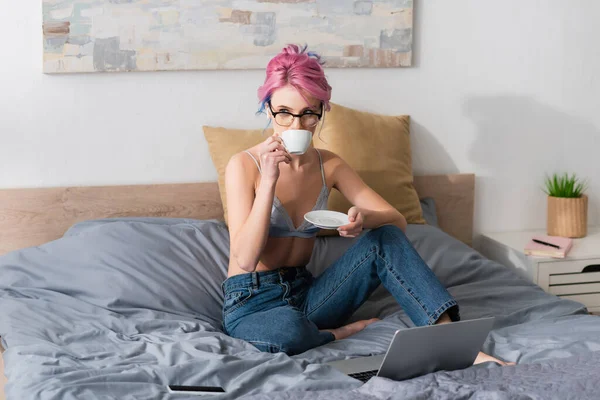 Jovem com cabelo rosa e fone de ouvido bebendo café perto de gadgets na cama — Fotografia de Stock