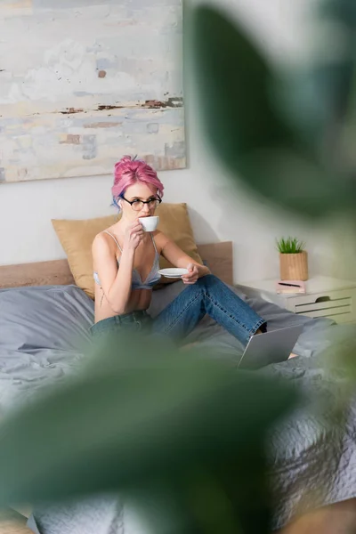 Mujer joven con el pelo rosa y auriculares beber café cerca de la computadora portátil en el dormitorio — Stock Photo