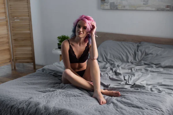 Tattooed and barefoot woman in dark underwear sitting in bedroom — Stock Photo