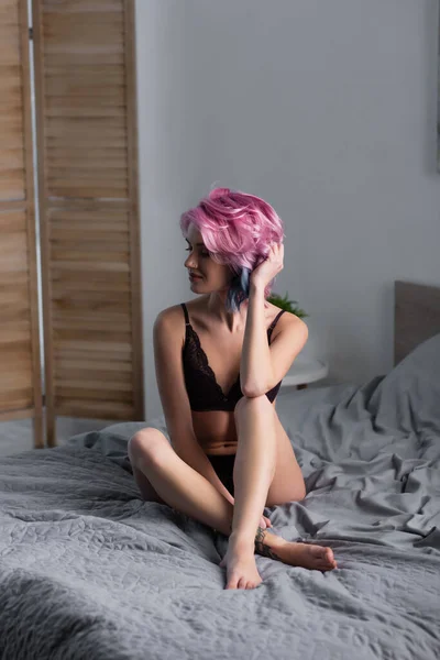 Joven mujer descalza en ropa interior oscura sentada en el dormitorio - foto de stock