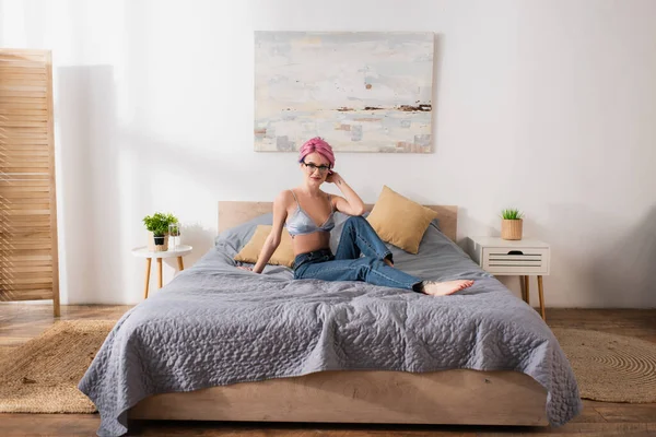 Bella giovane donna in jeans e reggiseno seduto sul letto in camera da letto moderna — Foto stock