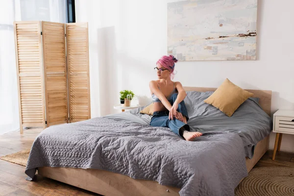 Barefoot young woman with dyed hair in jeans and bra sitting on bed in modern bedroom — Stock Photo