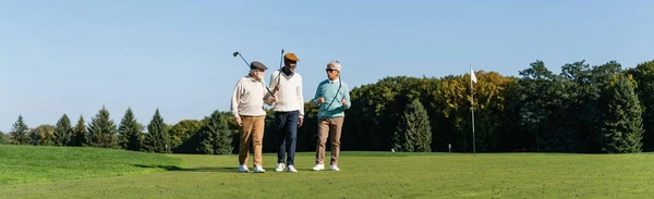Amis interracial seniors marchant avec des clubs de golf sur le terrain vert, bannière — Photo de stock