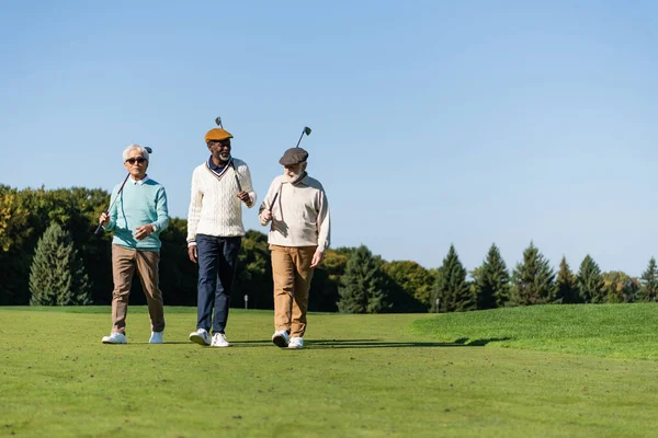 Senior interracial friends walking with golf clubs on green field — стоковое фото