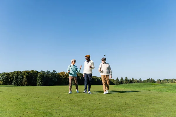 Amigos inter-raciais seniores andando com tacos de golfe em campo — Fotografia de Stock