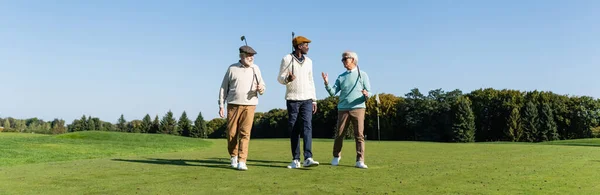 Senior interracial friends walking with golf clubs on field, banner — Stock Photo