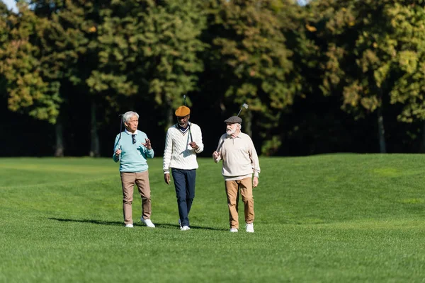 Interracial senior friends walking with golf clubs on field — Stock Photo