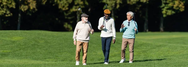 Amigos mayores interracial caminando con palos de golf en el campo, bandera - foto de stock