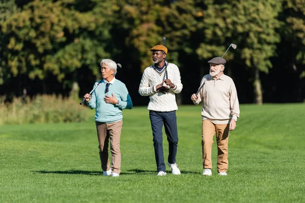 Homens seniores inter-raciais andando com tacos de golfe no campo verde — Fotografia de Stock