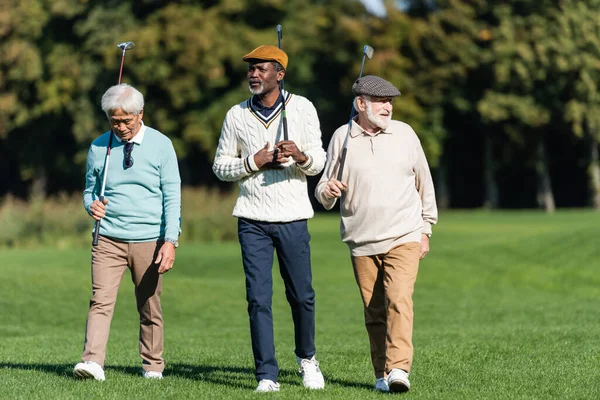 Amigos seniores inter-raciais andando com tacos de golfe fora — Fotografia de Stock