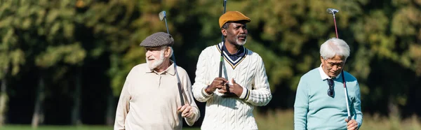 Amigos mayores interracial caminando con palos de golf afuera, pancarta - foto de stock