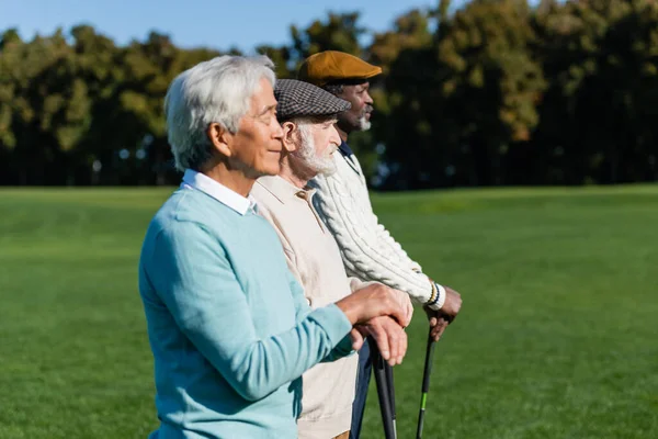 Vista laterale dell'uomo anziano in berretto piatto vicino agli amici interrazziali con mazze da golf — Foto stock