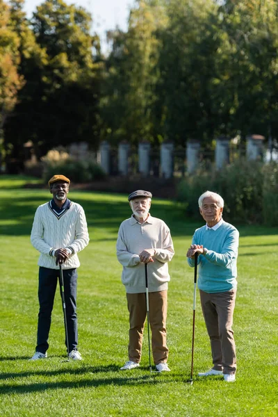Amigos mayores interracial de pie con palos de golf - foto de stock