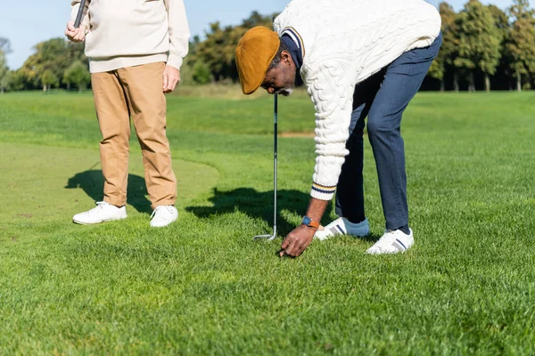 Afro-americano homem no flat cap colocando bola de golfe no gramado verde perto de amigo — Fotografia de Stock