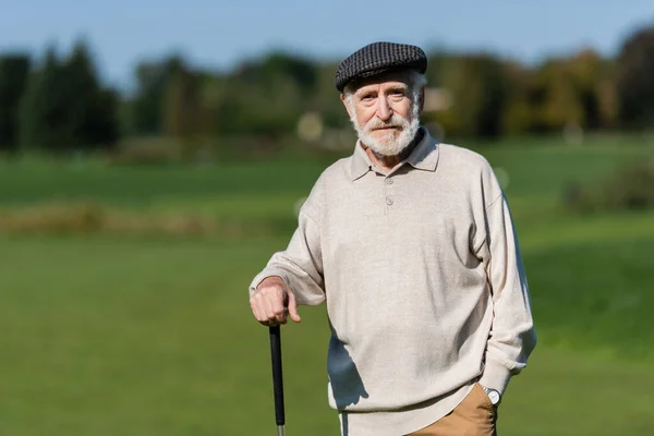 Uomo anziano ricco in berretto piatto in piedi con golf club — Foto stock
