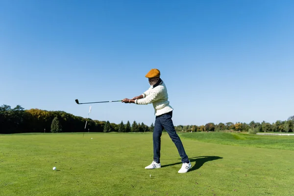 Comprimento total do homem americano africano em cap plana segurando clube de golfe e jogando no gramado verde — Fotografia de Stock