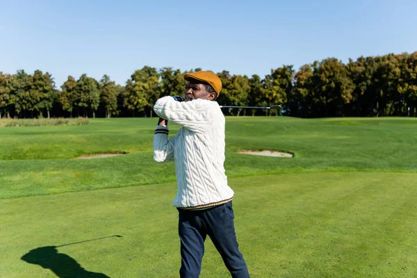 Middle aged african american man in flat cap holding golf club on green lawn — стоковое фото