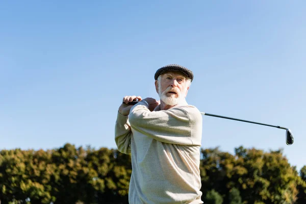 Vista de ángulo bajo del hombre mayor barbudo en la celebración de la tapa plana club de golf mientras juega al aire libre - foto de stock