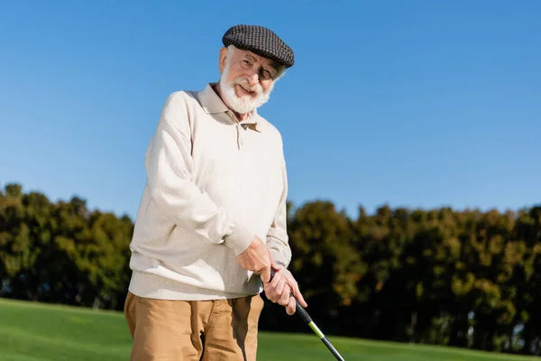 Happy senior man in flat cap holding golf club while playing outdoors — Stock Photo