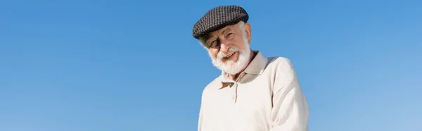 Heureux homme âgé en chapeau plat regardant la caméra contre le ciel bleu, bannière — Photo de stock
