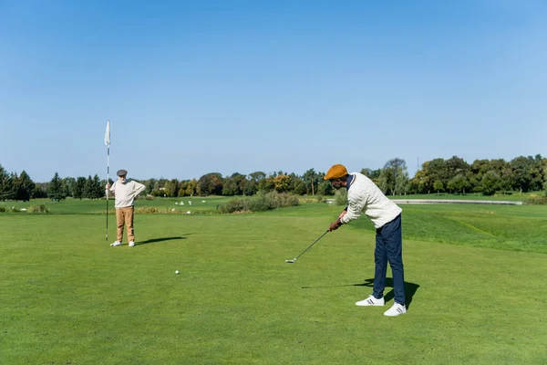 Senior hombre en plana gorra celebración bandera palo mientras africano americano amigo jugando golf - foto de stock