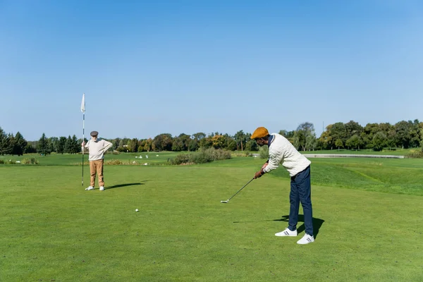 Homem sênior em cap plana segurando bandeira vara enquanto amigo afro-americano jogando golfe — Fotografia de Stock