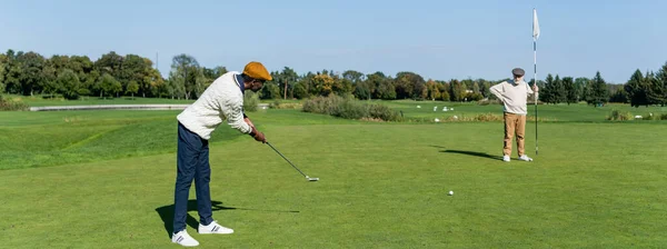 Senior man in flat cap holding flag stick while african american friend playing golf, banner — стоковое фото