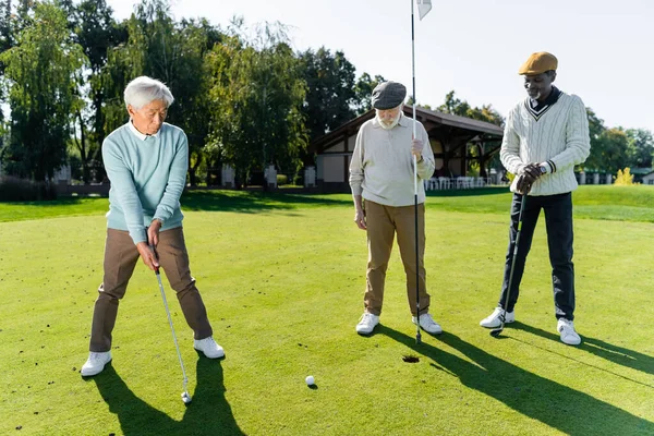 Senior man in flat cap holding flag stick near hole and multiethnic friends — Stock Photo