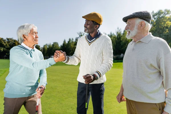 Sênior asiático e africano americano homens apertando as mãos enquanto segurando tacos de golfe perto alegre amigo — Fotografia de Stock