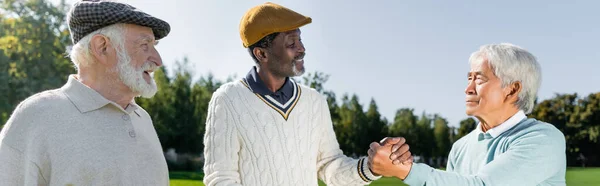 Senior asian and african american men shaking hands near cheerful friend, banner — Stock Photo