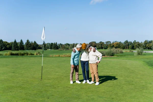 Senior interracial friends hugging near flag stick on green field — Stock Photo