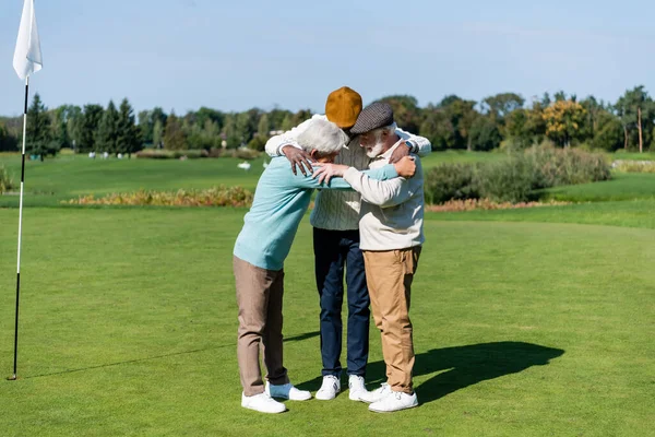 Senior multiethnic men hugging near flag stick on green field — Stock Photo