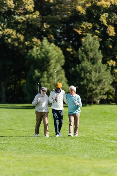 Senior multiethnic friends walking with golf clubs on green field near trees — стоковое фото
