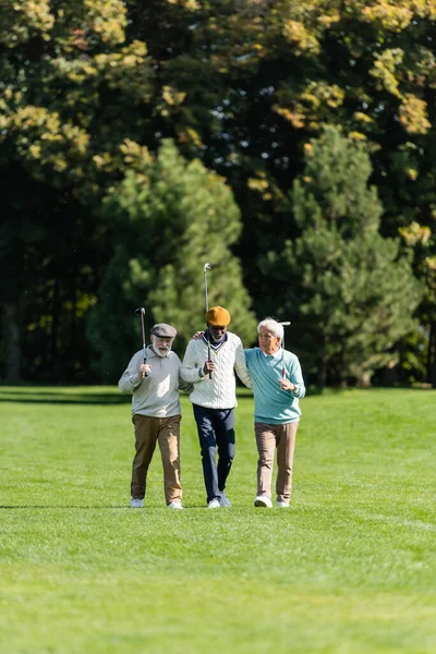 Senior multiethnic friends walking with golf clubs on green field near trees — Stock Photo