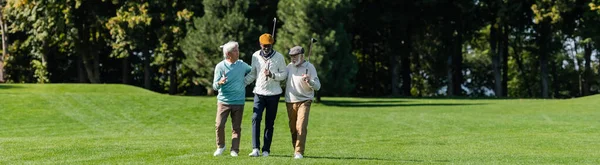 Hombres multiétnicos mayores caminando con palos de golf en el campo verde cerca de los árboles, pancarta - foto de stock