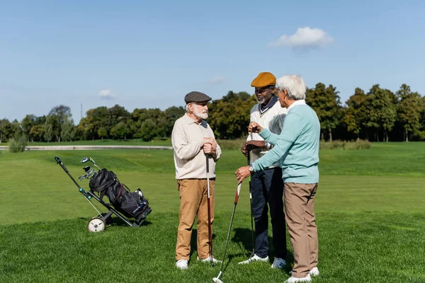 Senior multiethnic friends holding golf clubs and talking on field — Stock Photo