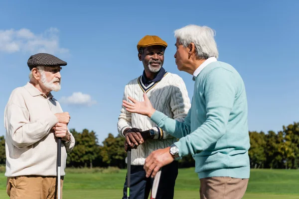 Senior interracial friends holding golf clubs and talking on field — Stock Photo