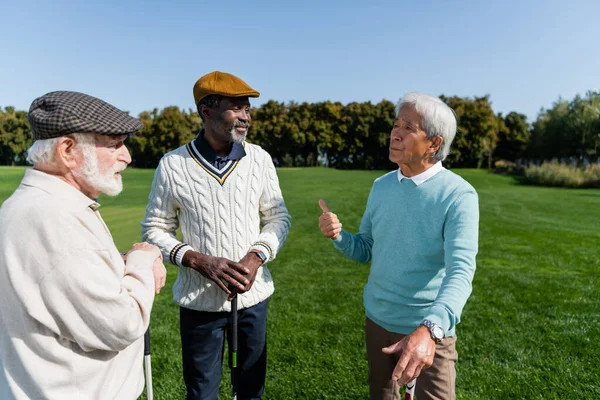 Senior asian man gesturing and talking with multiethnic friends on golf field — Stock Photo