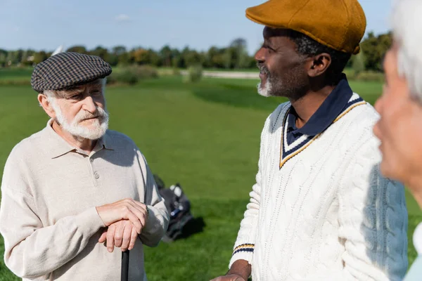 Idosos amigos multiétnicos olhando para o homem em boné plano no campo de golfe — Fotografia de Stock