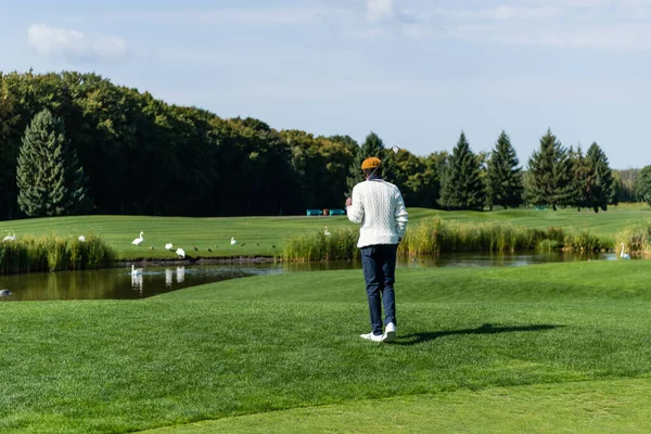 Вид ззаду афроамериканця, який тримає гольф-клуб і ходить на зеленому полі — стокове фото