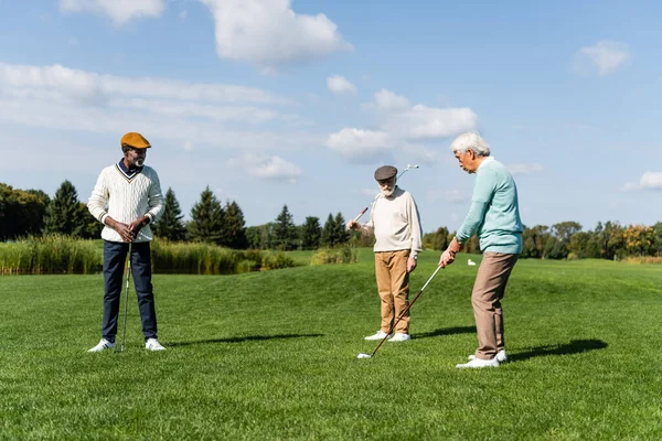 Счастливый африканский американец в плоской кепке смотрит на азиатского друга, играющего в гольф — стоковое фото